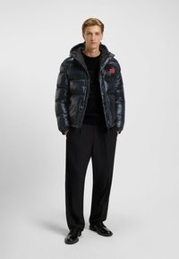 Giacca puffer blu navy con cappuccio, dotata di chiusura con zip, design trapuntato e patch del logo rosso. Indossata sopra un maglione nero e pantaloni.