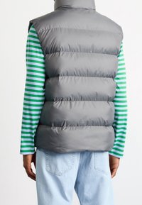 Grå pufferväst med hög krage, quiltat mönster, bärs över en grön och vit randig långärmad tröja. Avslappnade jeanstroser syns.