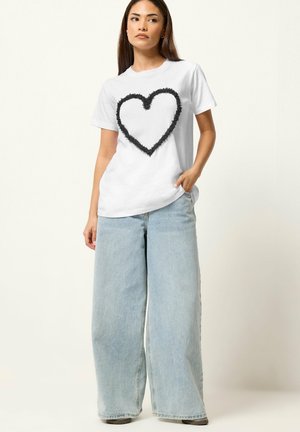 Frau steht mit der Hand in der Tasche, trägt ein weißes T-Shirt mit schwarzem flauschigem Herz und weit geschnittene hellblaue Jeans vor schlichter Hintergrund.