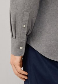 Camisa gris de manga larga con tejido texturizado; presenta puños abotonados y pliegues en la manga. Mano descansando sobre un pantalón azul marino.
