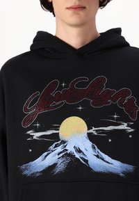 Svart hoodie med ett grafiskt tryck av ett berg och sol, accentuerad med röd text som säger "dina ljus" och glittrande rhinstenar.