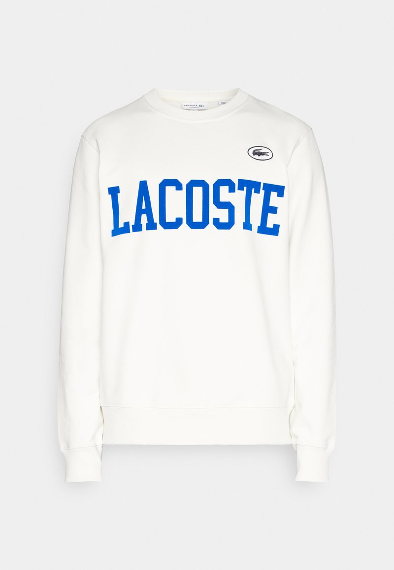 Felpa bianca in cotone, con un grande logo grafico "LACOSTE" blu sul petto e un piccolo emblema di coccodrillo verde sulla spalla.