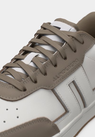 Κοντινό πλάνο λευκού και μπεζ αθλητικού παπουτσιού με μπεζ κορδόνια, τρύπες κοντά στη φτέρνα και ανάγλυφο όνομα μάρκας στην πλαϊνή πλευρά.