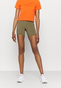 Camiseta naranja corta con pequeño logo negro, combinada con pantalones cortos deportivos verde oliva. El modelo lleva zapatillas de correr blancas. Diseño sencillo, estilo ajustado.