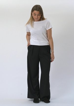 Vrouw in wit kort mouw overhemd en zwarte wijde pantalon met krijtstrepen staat met één hand in de zak, kijkt naar beneden tegen een effen achtergrond.