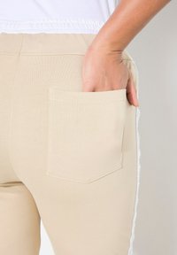 Béžové fitness kalhoty mají zadní kapsu čtvercového tvaru. Materiál je hladký a podél strany běží bílý akcent.