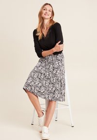 Schwarzes Langarmshirt mit Spitzenornament am Ausschnitt, kombiniert mit einem knielangen schwarz-weißen gemusterten Rock und weißen Turnschuhen.