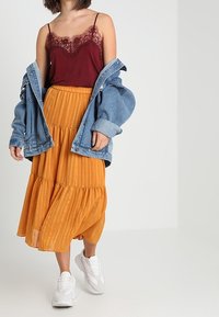 Camisola bordeaux con pizzo, abbinata a una gonna gialla senape a strati, giacca di jeans e sneakers bianche. Tessuto strutturato con righe.