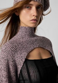 Ung kvinde med langt hår, iført en lavendel strikket beret, en matchende cropped turtleneck sweater og en sort gennemsigtig top.