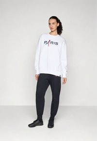 Witte sweatshirts met "PARIS" logo in marineblauw en rood; gecombineerd met zwarte joggers; model draagt zwarte sportschoenen. Gladde stoftextuur.