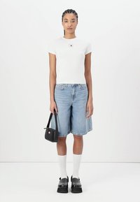 Camiseta ajustada blanca con un pequeño logo, pantalones cortos de mezclilla azules, bolso negro, calcetines blancos y zapatos negros gruesos. Fondo liso.