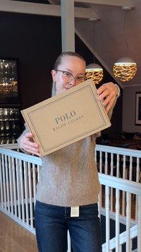 Kartonasta škatla z napisom "POLO RALPH LAUREN" v temnem besedilu na svetlo rjavi podlagi, ki jo drži oseba v bež puloverju.