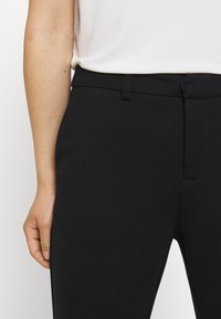 Primer plano de una persona que lleva pantalones ajustados negros y una blusa blanca metida por dentro, mostrando la cinturilla, los lazos del cinturón y el brazo izquierdo.