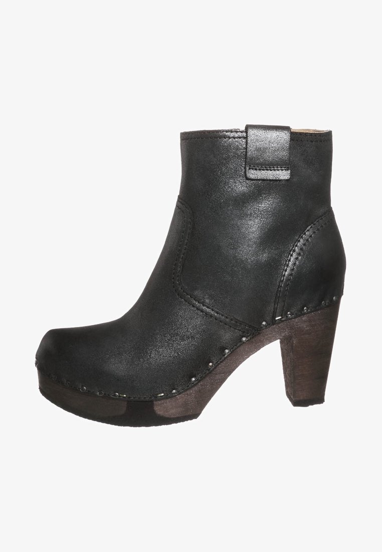 Bota de cuero negra hasta el tobillo con tacón alto. Presenta una punta redondeada, costuras visibles y pequeños acentos metálicos a lo largo de la suela.
