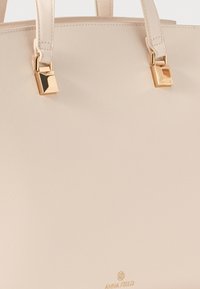 Lys beige håndtaske lavet af tekstureret syntetisk materiale, med guldfarvet hardware og dobbelte håndtag. Logo præsenteret i bunden.