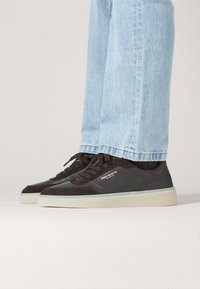 Ciemnobrązowe sneakersy z połączeniem gładkich i teksturowanych materiałów, z białą podeszwą i złotymi akcentami. Zestawione z jasnoniebieskimi dżinsami.