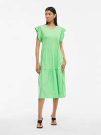 Zielona sukienka do kolan z miękkiego materiału, z krótkimi falbaniastymi rękawami i warstwowym krojem. Połączona z czarnymi sandałami.