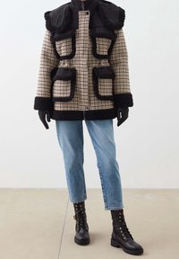 Persona vestida con un abrigo de cuadros con ribetes de piel de oveja negra, jeans azules, guantes de cuero negros y botas de combate negras con cordones, de pie sobre un suelo beige.