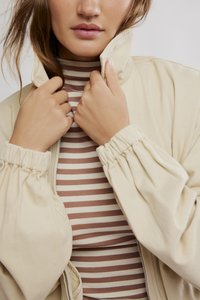 Beige Bomberjacke mit gerafften Ärmeln, offen getragen und darunter ein braun-creme gestreifter Rollkragenpullover. Glatte Stofftextur und lockere Passform.