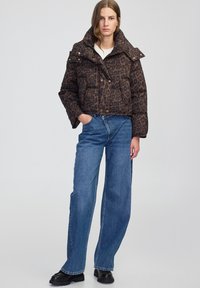 Bruine pufferjack met een luipaardpatroon, hoge kraag, drukknoopsluitingen en een gestructureerde stof; gedragen met brede blauwe jeans en zwarte schoenen.