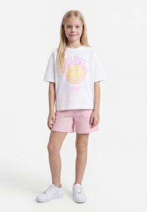 Junges Mädchen steht, trägt ein weißes T-Shirt mit einem gelben Smiley und pinkem Text, pinke Shorts und weiße Turnschuhe vor einem schlichten weißen Hintergrund.