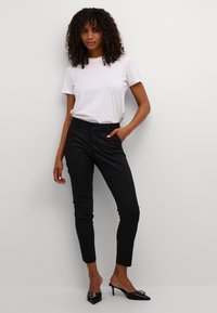 Pantalon noir tailleur, design ajusté, texture lisse, avec poches latérales. Associé à un t-shirt blanc uni à manches courtes et des mules à talons hauts.