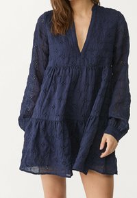 Robe bleu marine en tissu texturé avec un décolleté en V profond, des manches longues et des motifs en œillets floraux, présentant un bas évasé et en couches.
