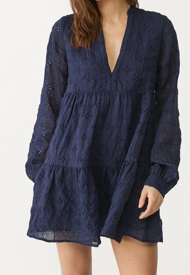 Robe bleu marine en tissu texturé avec un décolleté en V profond, des manches longues et des motifs en œillets floraux, présentant un bas évasé et en couches.