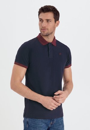 Man die een donkerblauw poloshirt draagt met een bordeauxrode kraag en mouwranden, staat met gevouwen handen en kijkt naar links.