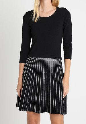 Robe noire avec un haut ajusté et une jupe plissée. La jupe présente de fines rayures verticales blanches. Fabriquée en tissu doux avec un col rond.