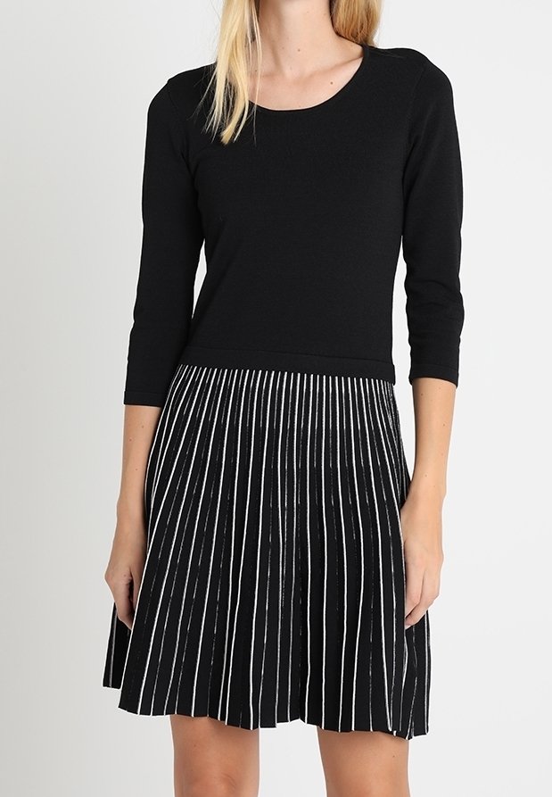 Robe noire avec un haut ajusté et une jupe plissée. La jupe présente de fines rayures verticales blanches. Fabriquée en tissu doux avec un col rond.