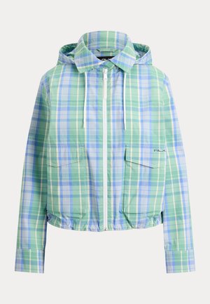 Femme debout portant une veste zippée à carreaux bleu-vert, un short blanc, des chaussettes blanches et des chaussures blanches à lacets sur un fond neutre.