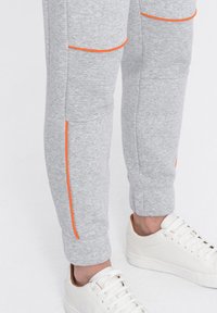 Graue Sport-Jogginghosen mit orangefarbenen Akzentnähten und tapered Bündchen, kombiniert mit weißen Sneakers mit hellbraunen Fersenakzenten.