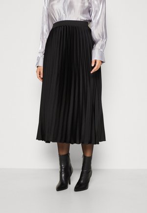 Frau trägt einen schwarzen plissierten Midirock, transparente schwarze Strumpfhosen, schwarze Stiefeletten mit Absatz und eine langärmelige silberne Satinbluse.