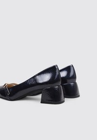 Des talons en cuir verni noir avec un bout arrondi, présentant une surface texturée et un petit talon bloc géométrique. Accentué d'un petit détail métallique.