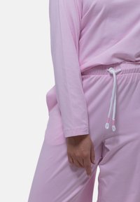 Ljust rosa långärmad tröja i kombination med matchande byxor. Byxorna har resårmidja och vita snördetaljer. Mjukt, slätt tyg.