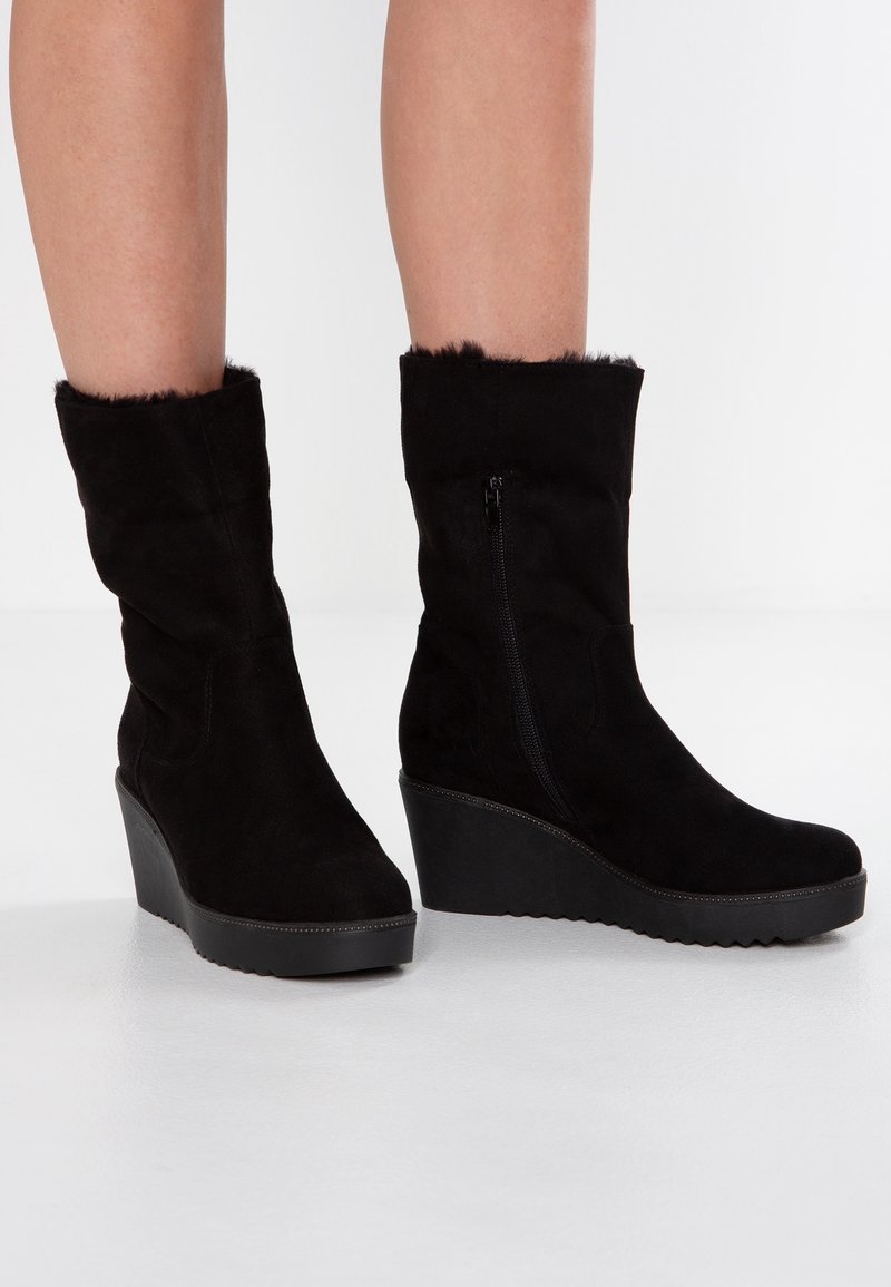 Bottes compensées en suède noir à cheville avec fermeture éclair latérale et bordure en fourrure, portées sur des jambes nues sur un fond blanc uni.