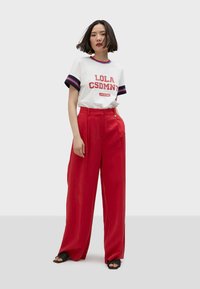 White graphic t-shirt with "LOLA CSDMN" in red, paired with high-waisted red wide-leg trousers and black open-toed sandals.