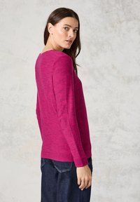 Helle fuchsia Langarmstrickjacke mit strukturiertem Rippenmuster, runder Halsauschnitt und lockerer Passform. Kombiniert mit dunklen Jeans.