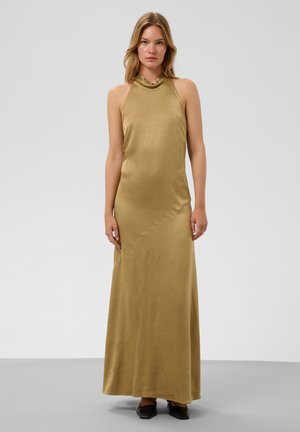 Femme portant une robe maxi en satin vert olive sans manches avec un col montant et des chaussures noires à bout pointu, debout devant un fond uni.