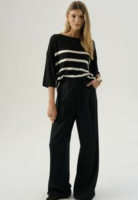 Donna che indossa un top leggero a righe bianche e nere, pantaloni neri a gamba larga, bracciale d'oro e anelli, in piedi di fronte a uno sfondo semplice.
