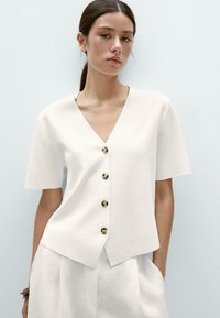 Blouse blanche à manches courtes avec un col en V et cinq boutons marron sur le devant. Tissu doux, coupe décontractée et ourlet légèrement asymétrique.