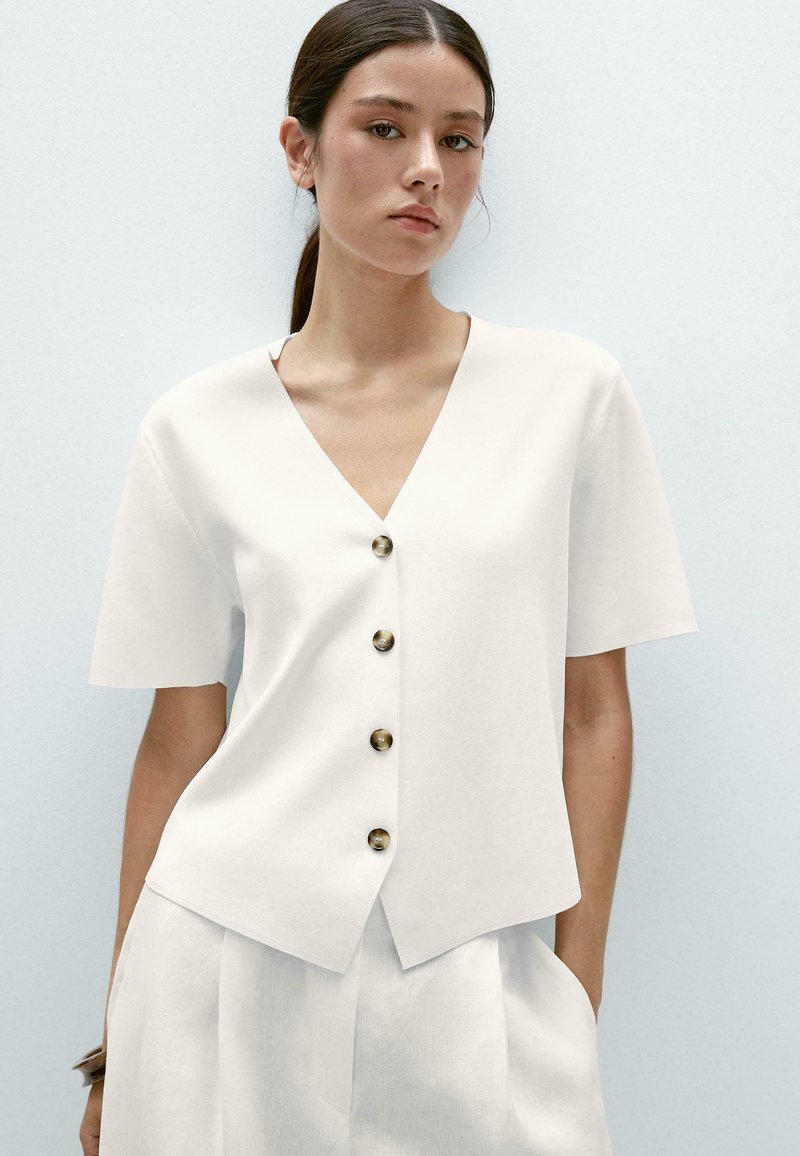 Blouse blanche à manches courtes avec un col en V et cinq boutons marron sur le devant. Tissu doux, coupe décontractée et ourlet légèrement asymétrique.