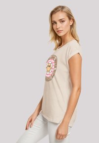 Beige cotton t-shirt with short sleeves, featuring a graphic of a smiling flower and circular text in black and pink. Worn with white jeans.