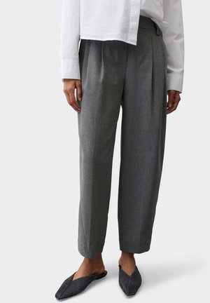 Person trägt graue plissierte Hose, ein weißes Langarmhemd und schwarze spitze flache Schuhe, steht auf hellem Hintergrund.