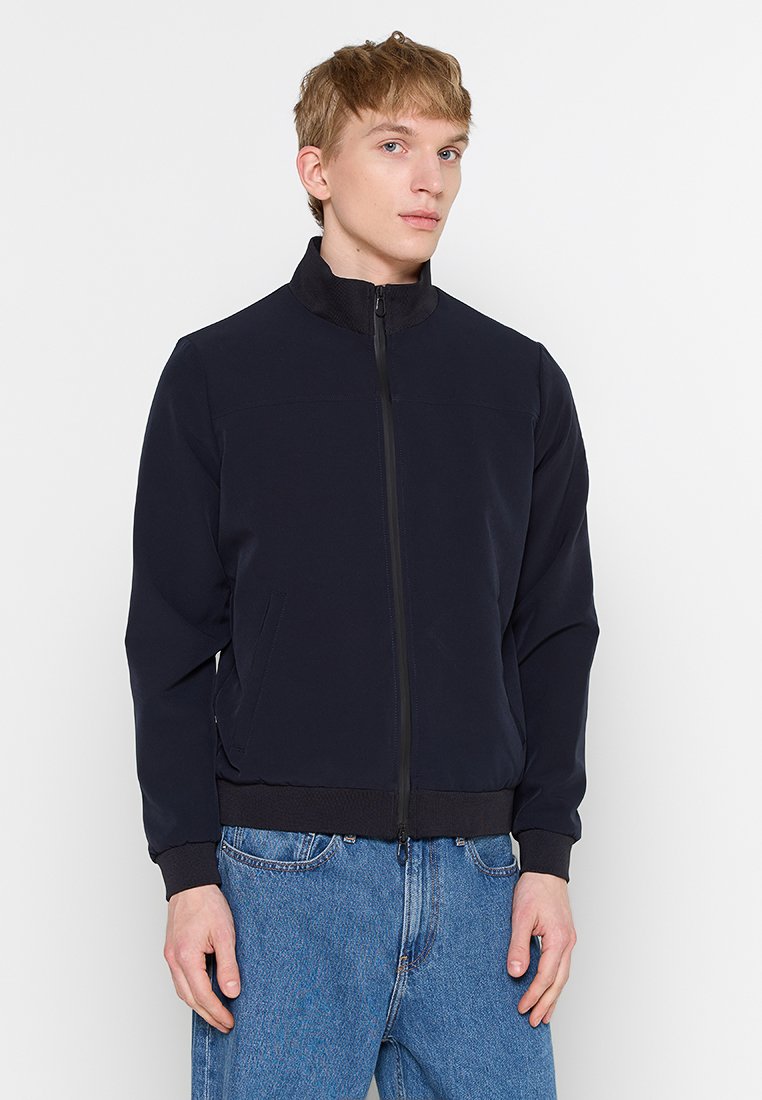 Joven vistiendo una chaqueta negra con cremallera, con puños y cuello acanalados, combinada con jeans azules, de pie contra un fondo blanco.