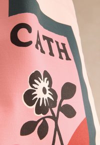 Drobės krepšys su gėlių raštu juodai baltame fone ant rožinės spalvos fono, kuriame ryškiai matomas užrašas „CATH“ storu juodu šriftu.
