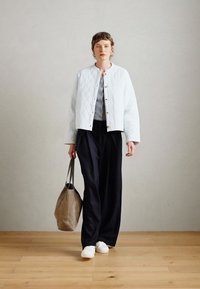 Casaco branco acolchoado com gola redonda e botões dourados, blusa cinza, calças pretas largas, sapatos brancos, a segurar uma bolsa bege.