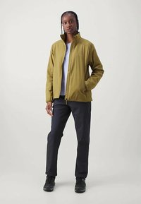 Άτομο που στέκεται φορώντας ένα ελαιοπράσινο φερμουάρ μπουφάν, λευκό πουκάμισο, μαύρο παντελόνι και μαύρα αθλητικά παπούτσια σε ένα απλό φόντο.