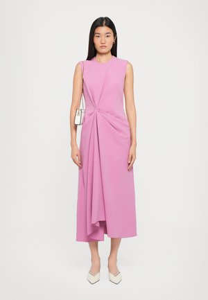 Pink sleeveless dress with a twist design at the waist, flowing fabric, and an asymmetrical hem. Model holding a small white handbag.